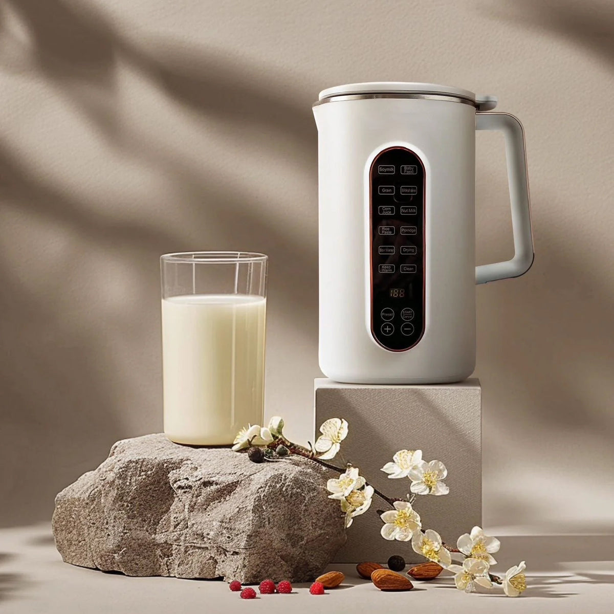 White AmonNusy Nut Milk Maker frother on a stone block with a glass of milk and flowers on a neutral background