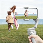 A handheld Auralink Bytech Gimbal in the foreground holding a phone with a video call interface, while two children run across a grassy field toward the ocean in the background. A hand is visible holding the gimbal.