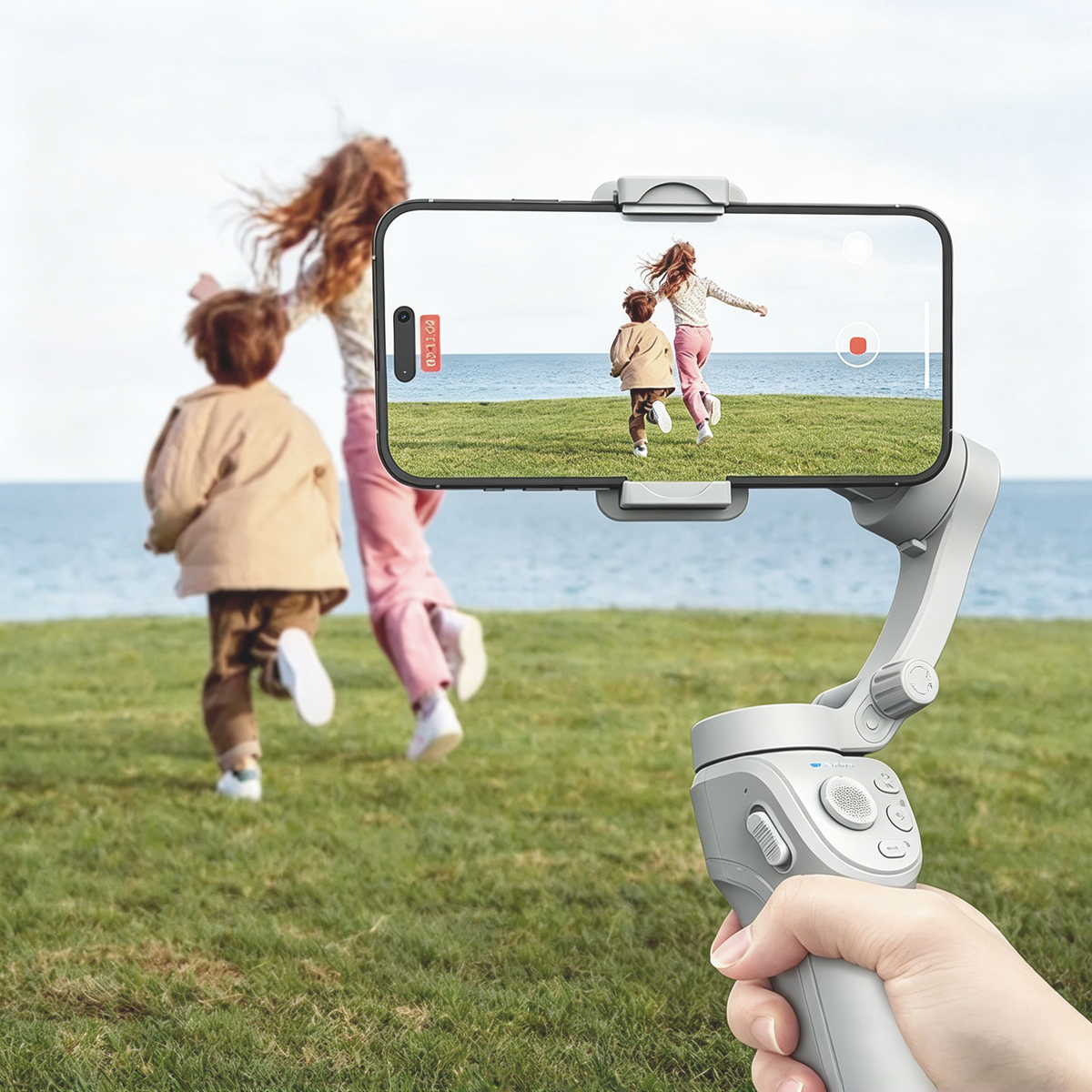 A handheld Auralink Bytech Gimbal in the foreground holding a phone with a video call interface, while two children run across a grassy field toward the ocean in the background. A hand is visible holding the gimbal.