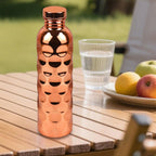 A CopperSyp Copper Water Bottle with a textured honeycomb pattern and a matching cap, standing on a wooden table outdoors next to a glass of water and fruit.