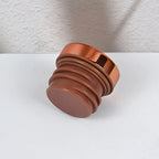 A metallic copper-colored cylindrical knob or decorative hardware piece with a ribbed base, shown on a white surface against a textured white wall.