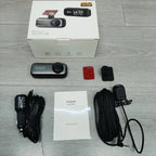 A boxed DashyLynk 4K Wireless Dashcam kit laid out on a light gray wood surface, including the retail box, a small black dash camera, mounting accessories, a car charger cable, a mounting bracket cable, a red mounting piece, and an instruction booklet.