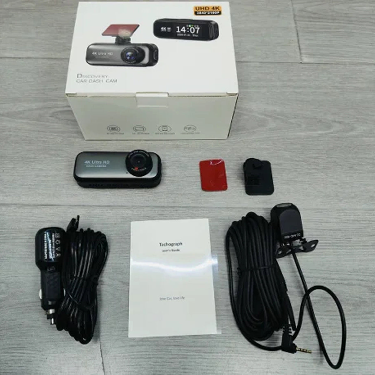 A boxed DashyLynk 4K Wireless Dashcam kit laid out on a light gray wood surface, including the retail box, a small black dash camera, mounting accessories, a car charger cable, a mounting bracket cable, a red mounting piece, and an instruction booklet.