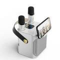 A white portable FolbGent Karaoke Machine with two built-in microphones, a black carrying handle with a gold button, and a built-in phone holder displaying a video call.