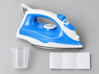 A blue and white Hilio Iron with a white cord, shown alongside a clear plastic measuring cup and a folded white fabric piece on a light gray background.