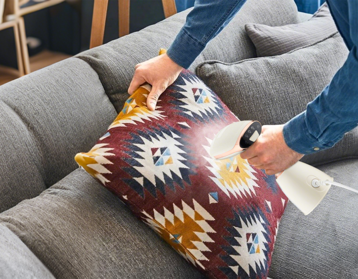 Person using a Krevont Steamer on a decorative pillow on a gray couch.