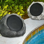 Two rock-shaped speakers, one dark gray and one silver, placed beside a swimming pool with green foliage in the background.
