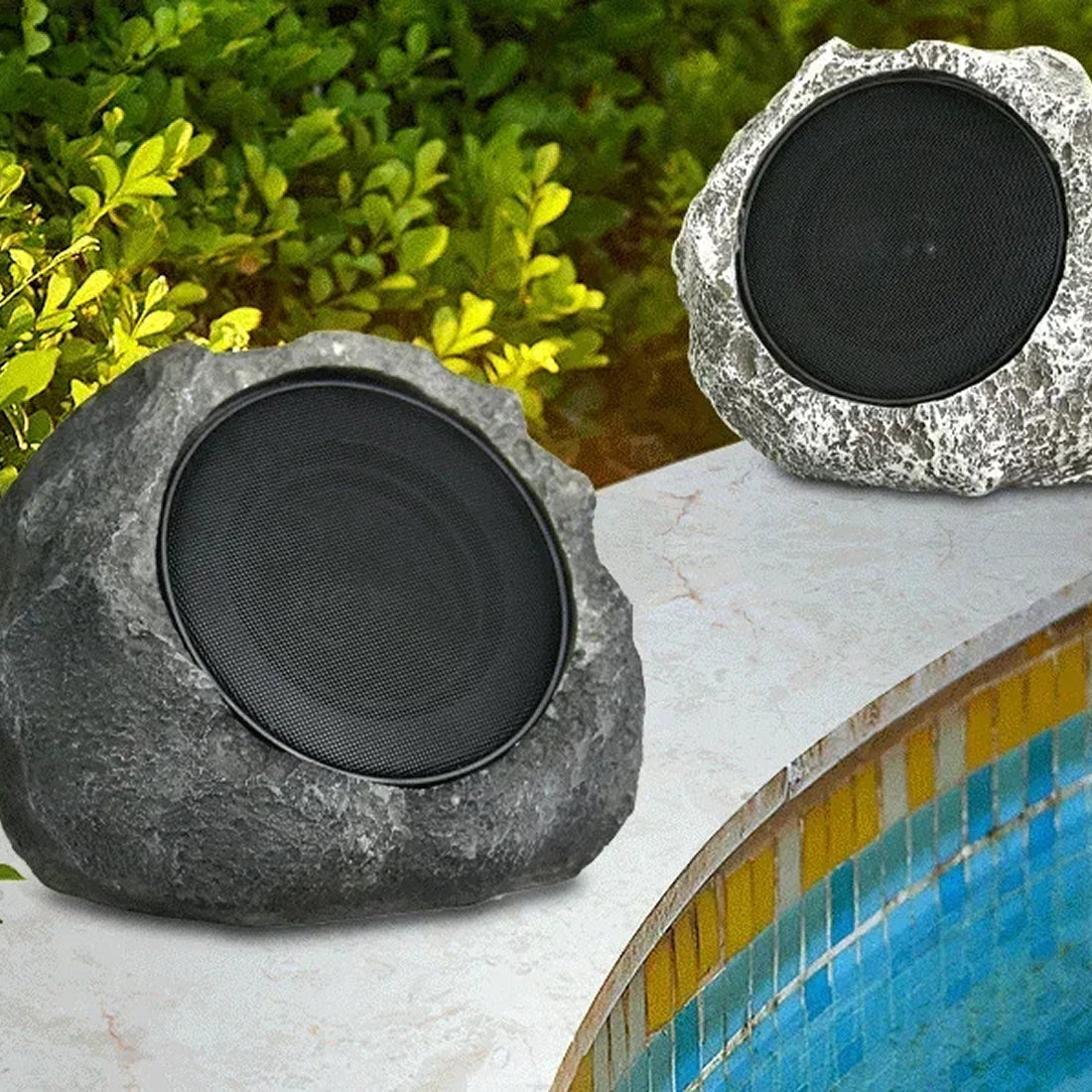 Two rock-shaped speakers, one dark gray and one silver, placed beside a swimming pool with green foliage in the background.