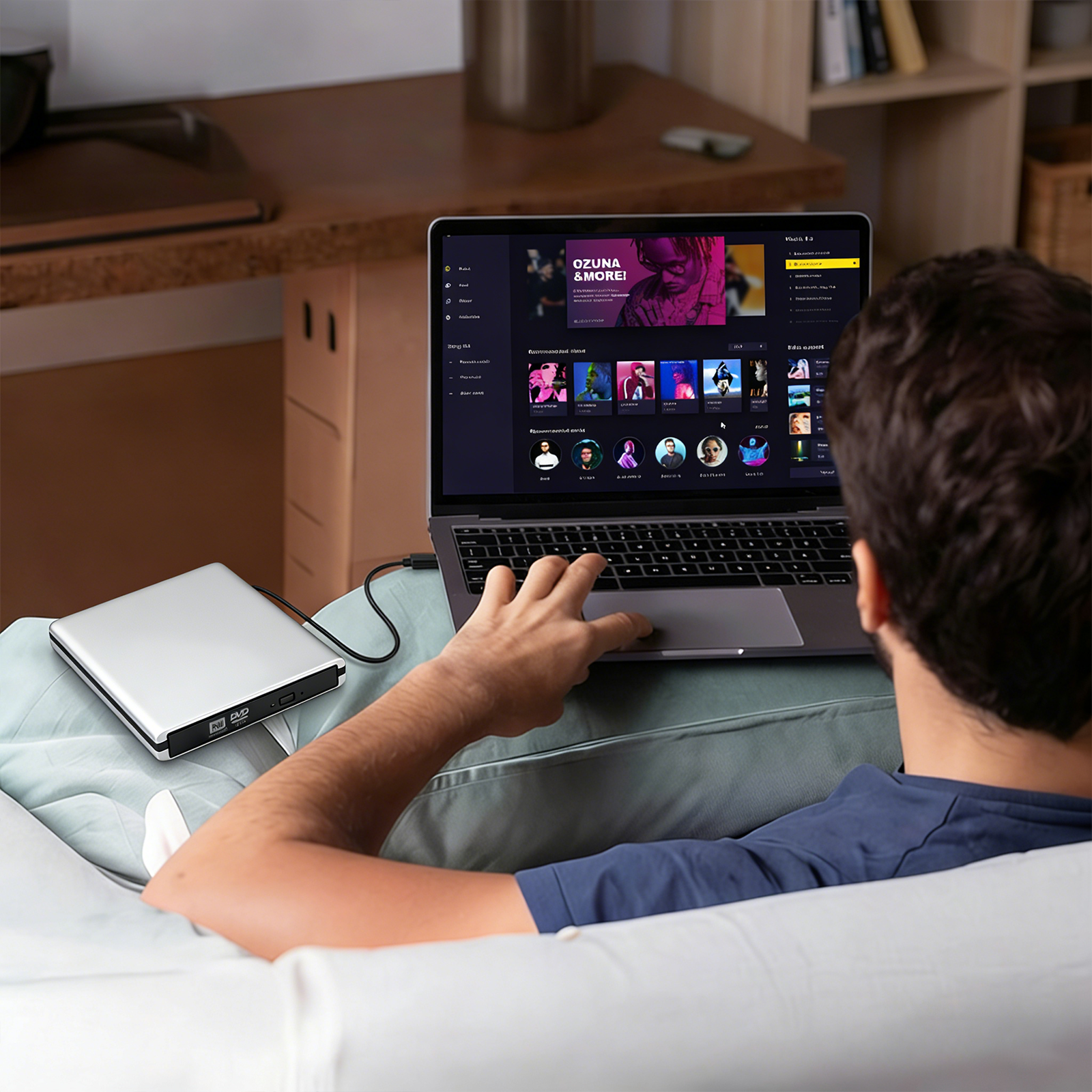 A man reclining on a couch uses a black laptop on his lap, with a silver external hard drive connected via cable on the couch beside him in a home office setting.