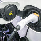 A close-up of an electric vehicle Portable EV Charger for Type 2 Cars connector plugged into a charging port on a white EV car, with raindrops visible on the connector and water droplets in the air.