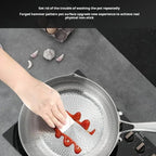 A close-up of a hand using a white cleaning cloth to wipe red sauce from the inside of a hammered stainless steel frying pan on a black stovetop. Garlic cloves and an egg carton are visible on a gray countertop, with a black text banner at the top describing the non-stick coating.